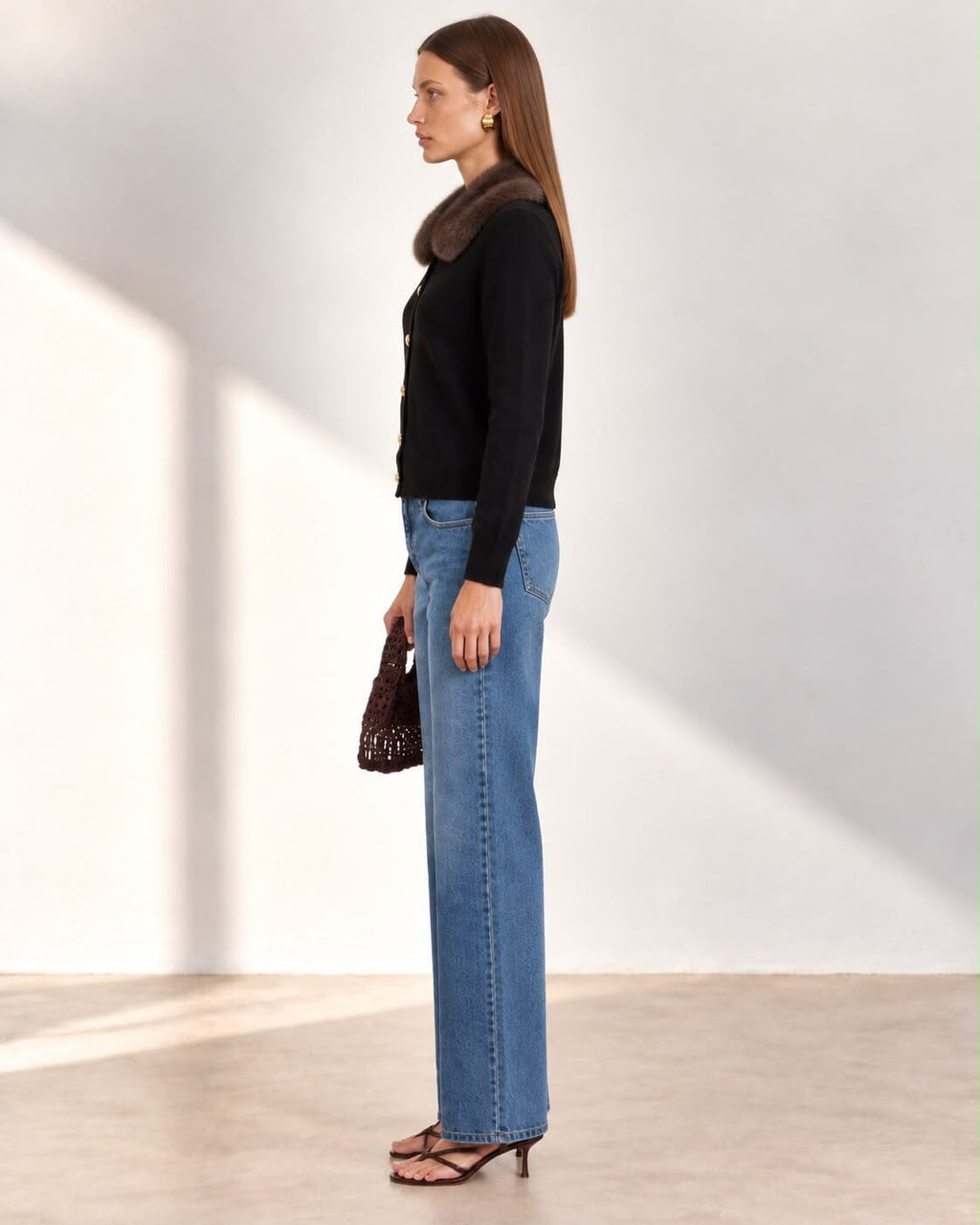 A woman stands in a studio wearing a black long-sleeve cardigan with a large brown fur-like collar and gold buttons, paired with blue straight-leg jeans, brown sandals, and holding a small brown textured handbag.