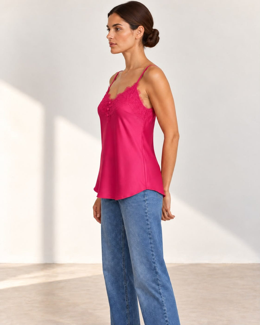 A woman modeling a bright pink satin camisole top with lace trim and blue jeans in a minimalist studio setting.
