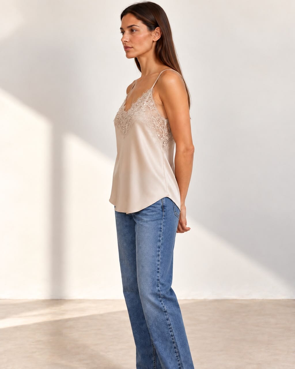 A woman modeling a light beige satin camisole top with lace detailing on the cups, paired with blue jeans, standing against a minimalist off-white wall with soft shadows.