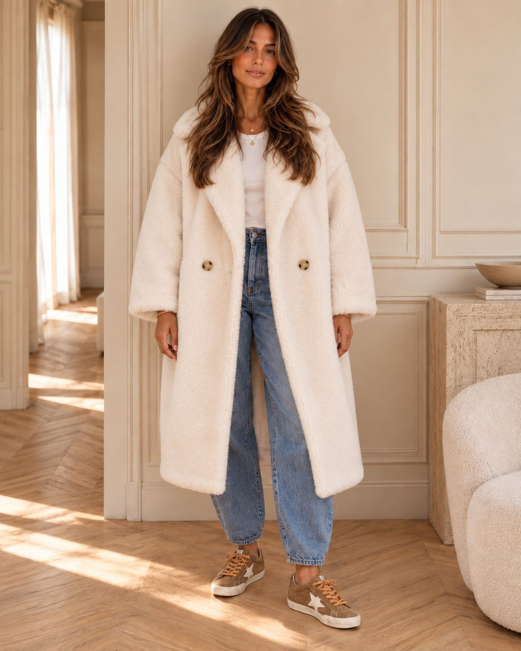 A woman wearing a long cream faux fur coat, blue jeans, and brown sneakers stands in a sunlit room with beige walls and wood floors.