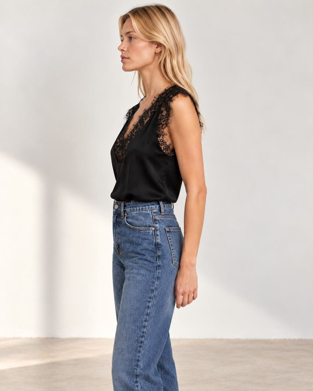 A woman modeling a black lace-trimmed satin top with blue jeans, standing against a plain light gray studio wall.