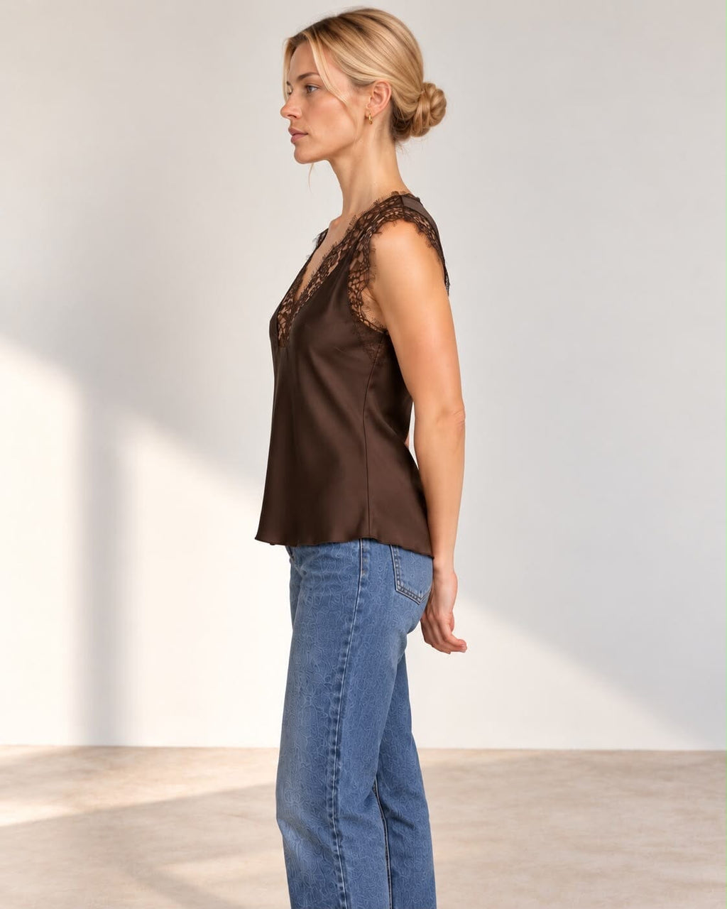 A woman modeling a sleeveless dark brown satin top with a lace V-neckline, paired with blue jeans, standing against a neutral studio wall.