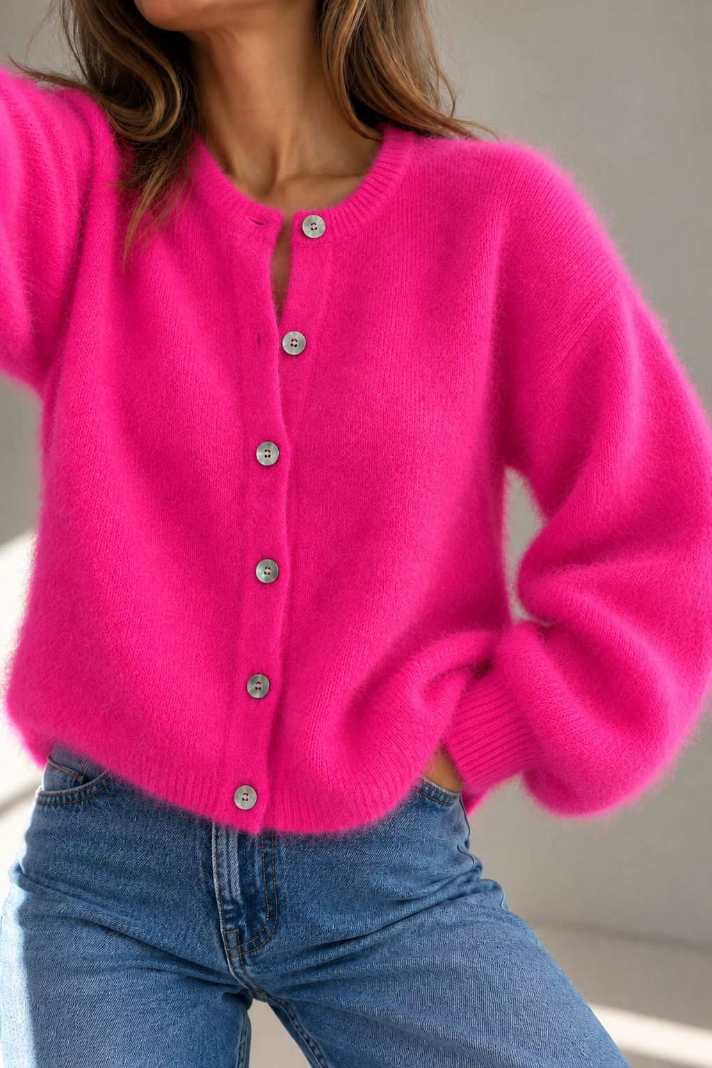 A woman wearing a bright hot pink fuzzy cardigan over a white tank top and light blue jeans, holding a beige shoulder bag and a magazine against a gray textured wall.