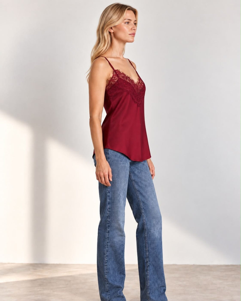 A woman modeling a burgundy satin camisole top with lace trim and blue jeans against a plain light gray studio background.