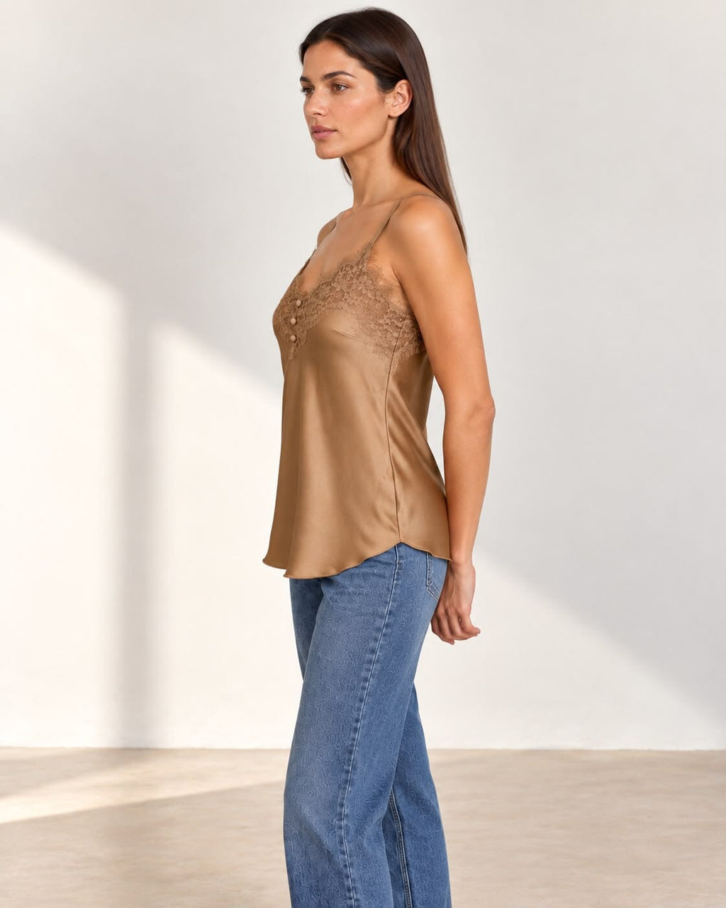 A woman modeling a beige satin camisole top with lace detailing on the bust, paired with blue jeans, standing against a minimalist off-white wall.