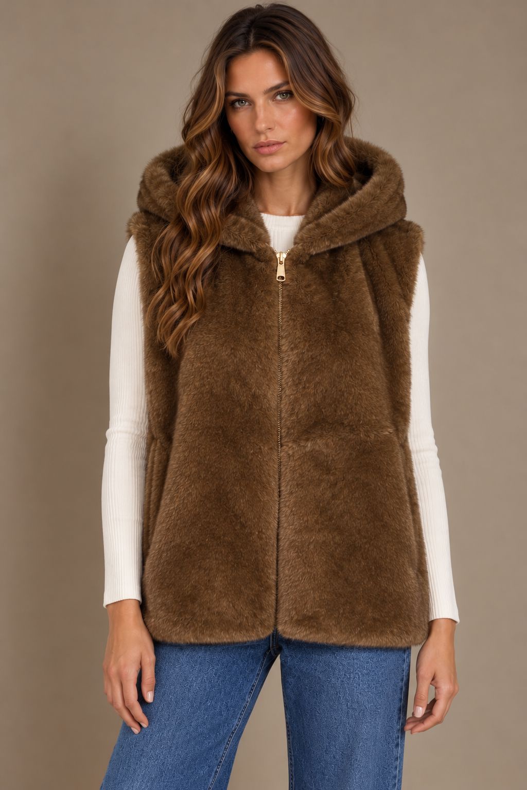 A woman wearing a brown faux fur hooded vest over a white ribbed long-sleeve top and blue jeans, photographed against a plain beige studio background.