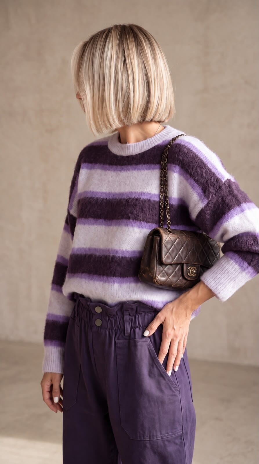 A woman wearing a fuzzy purple and white striped crewneck sweater paired with high-waisted dark purple pants, carrying a brown shoulder bag against a textured beige wall.