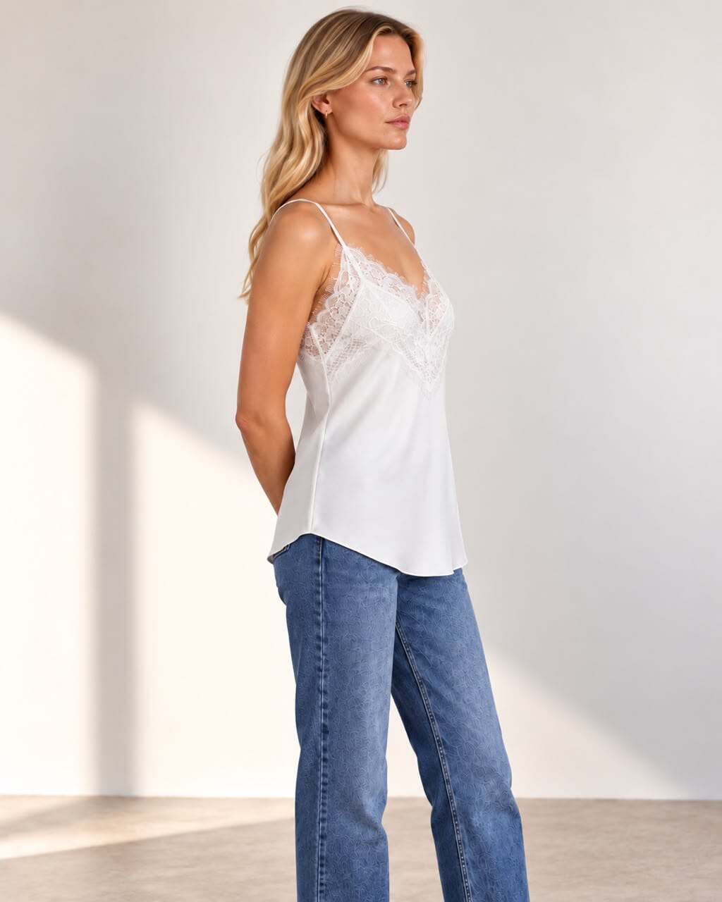 A woman modeling a white satin camisole top with delicate lace underbust detailing, paired with blue jeans, set against a minimalist off-white studio background with soft shadows.